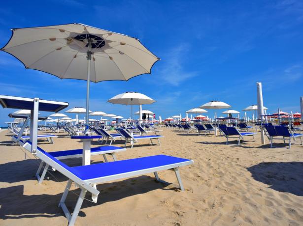 Spiaggia con lettini blu e ombrelloni bianchi sotto cielo sereno.