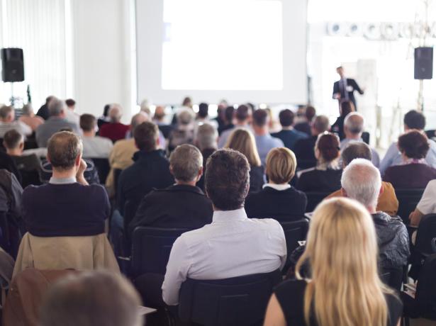 Persone in una conferenza, ascoltano un relatore davanti a uno schermo.